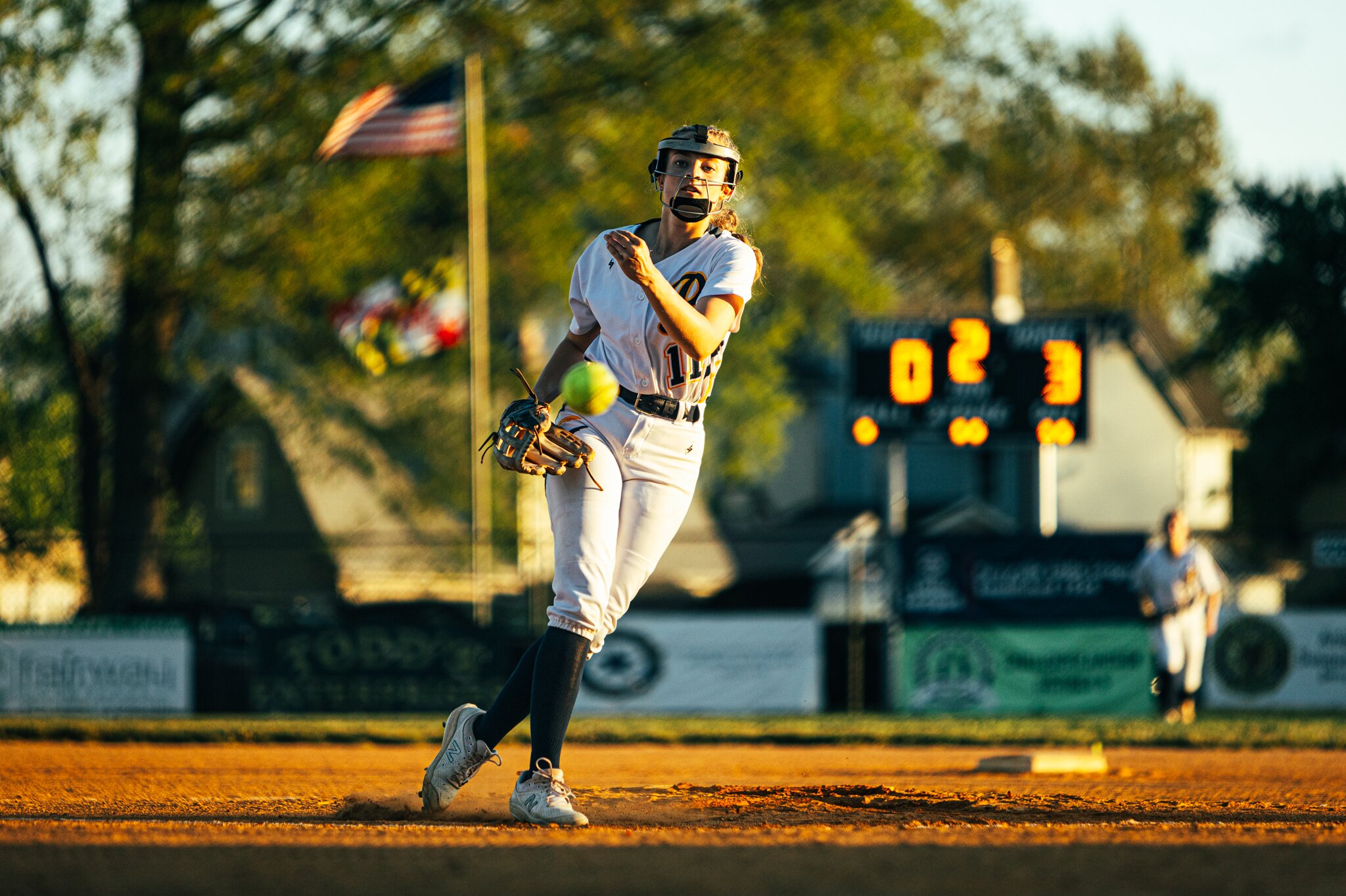 Softball pitcher releasing the ball in golden evening light with the flag and scoreboard behind her