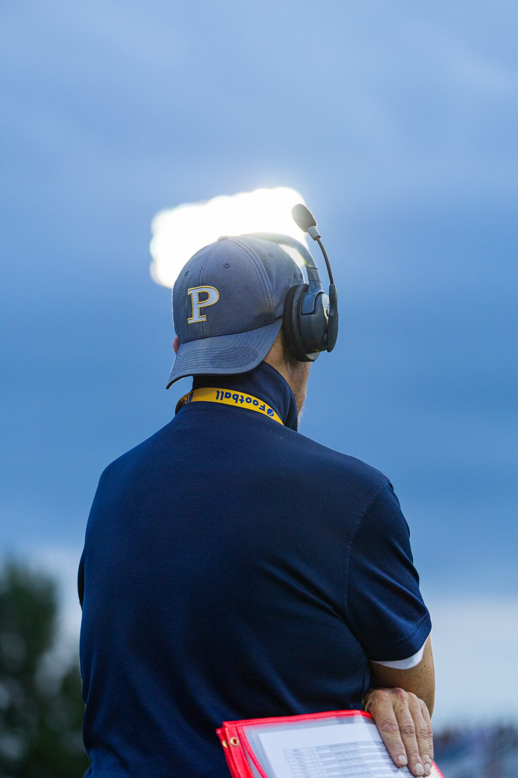 Coach seen from behind in a headset watching the field at dusk with a stadium light glowing above him