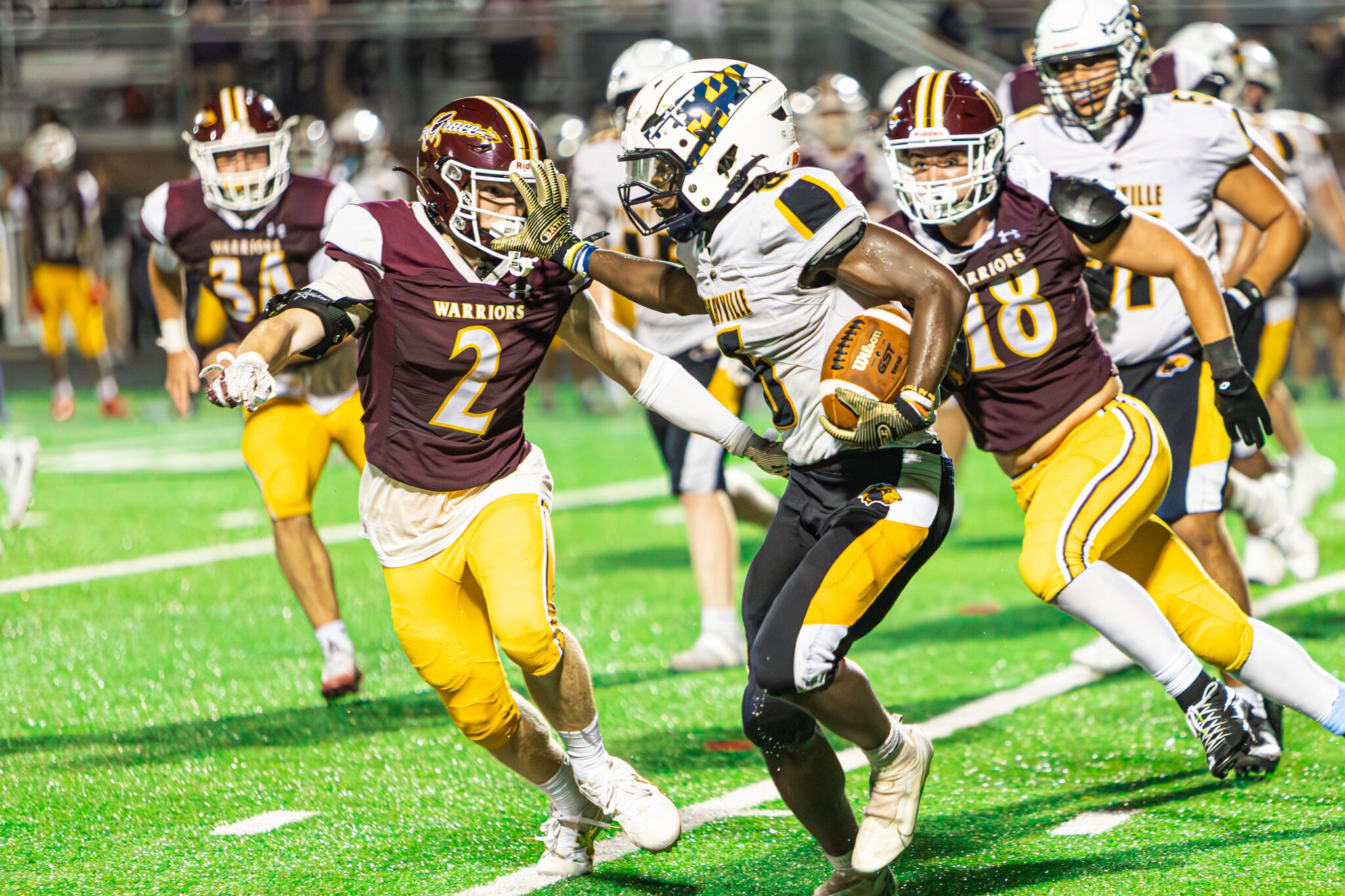 Football ball carrier in a white and gold jersey breaking through two defenders under stadium lights at night