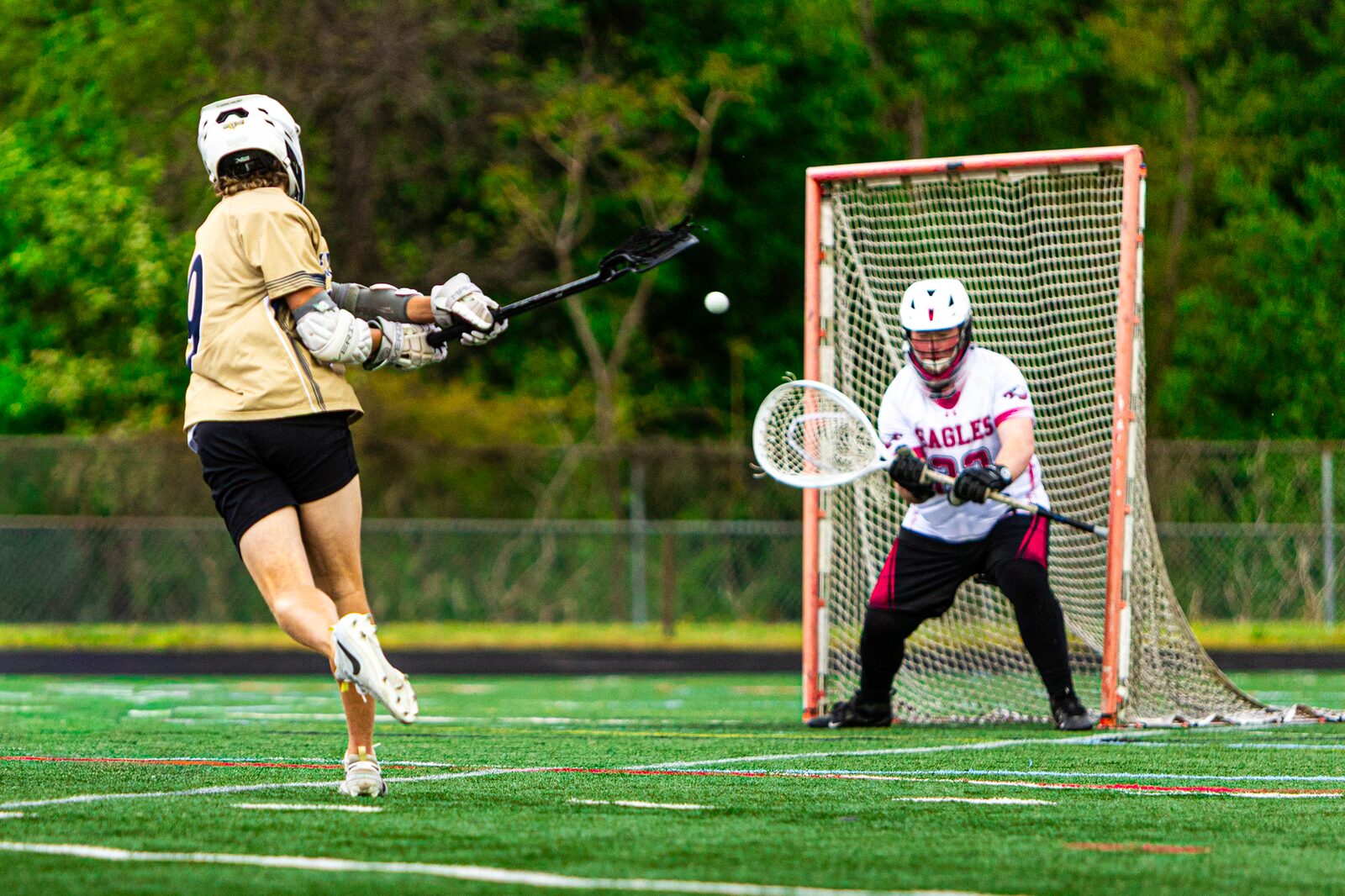 Lacrosse attacker firing a shot on goal as the keeper sets in the crease and the ball hangs in the air